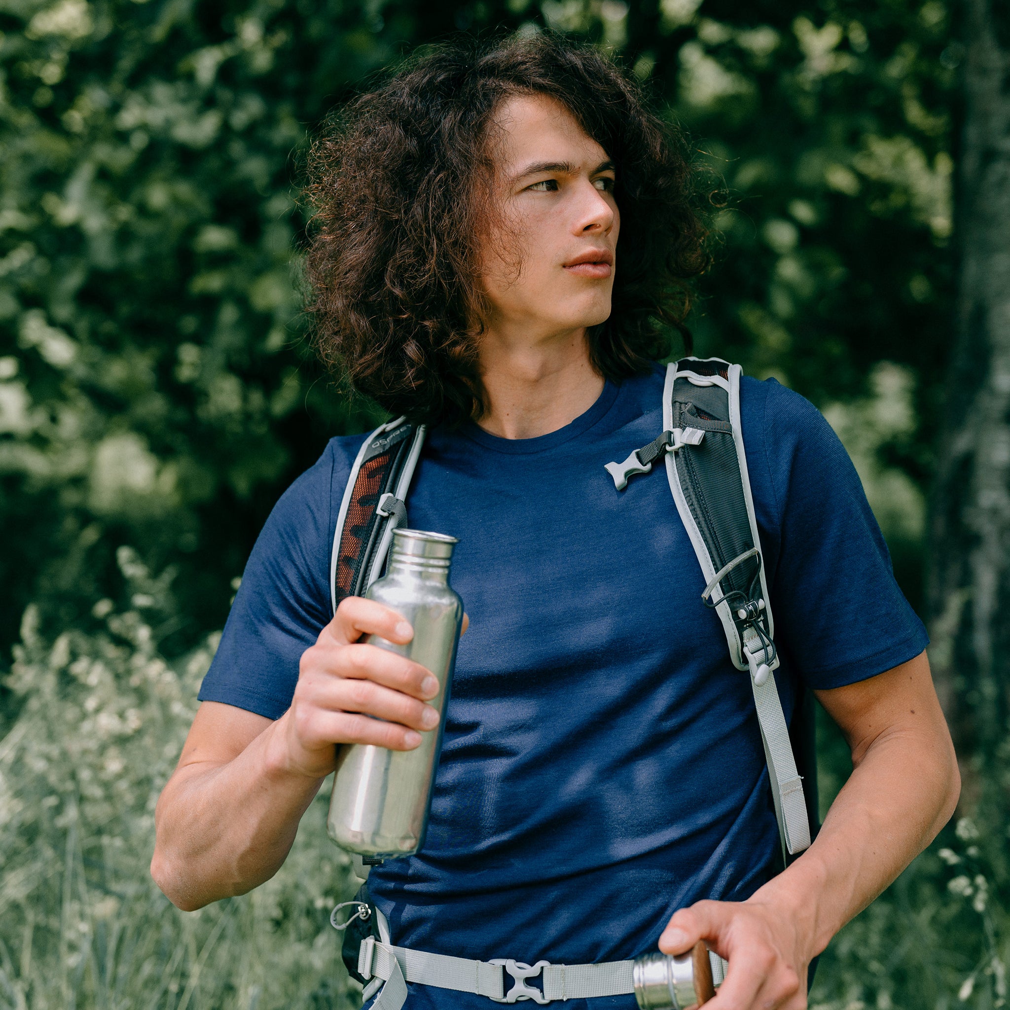 Model steht im blauen Shirt von Tom Fyfe aus Merinowolle vor einem Baum und trinkt aus der Flasche