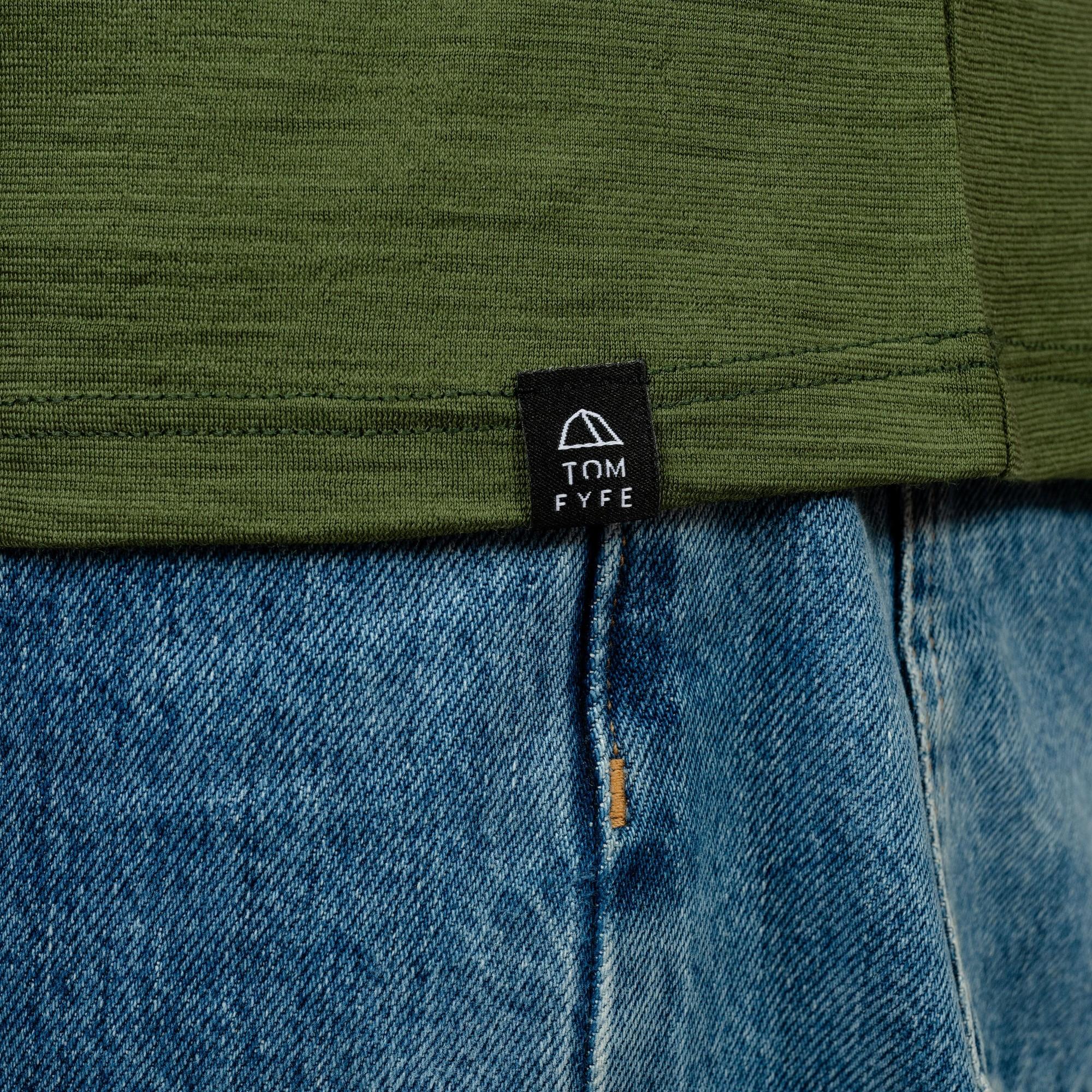 Model mit Merino Herren Shirt mit angenaehtem Stofflabel mit Logo von TOM FYFE in Waldgrün