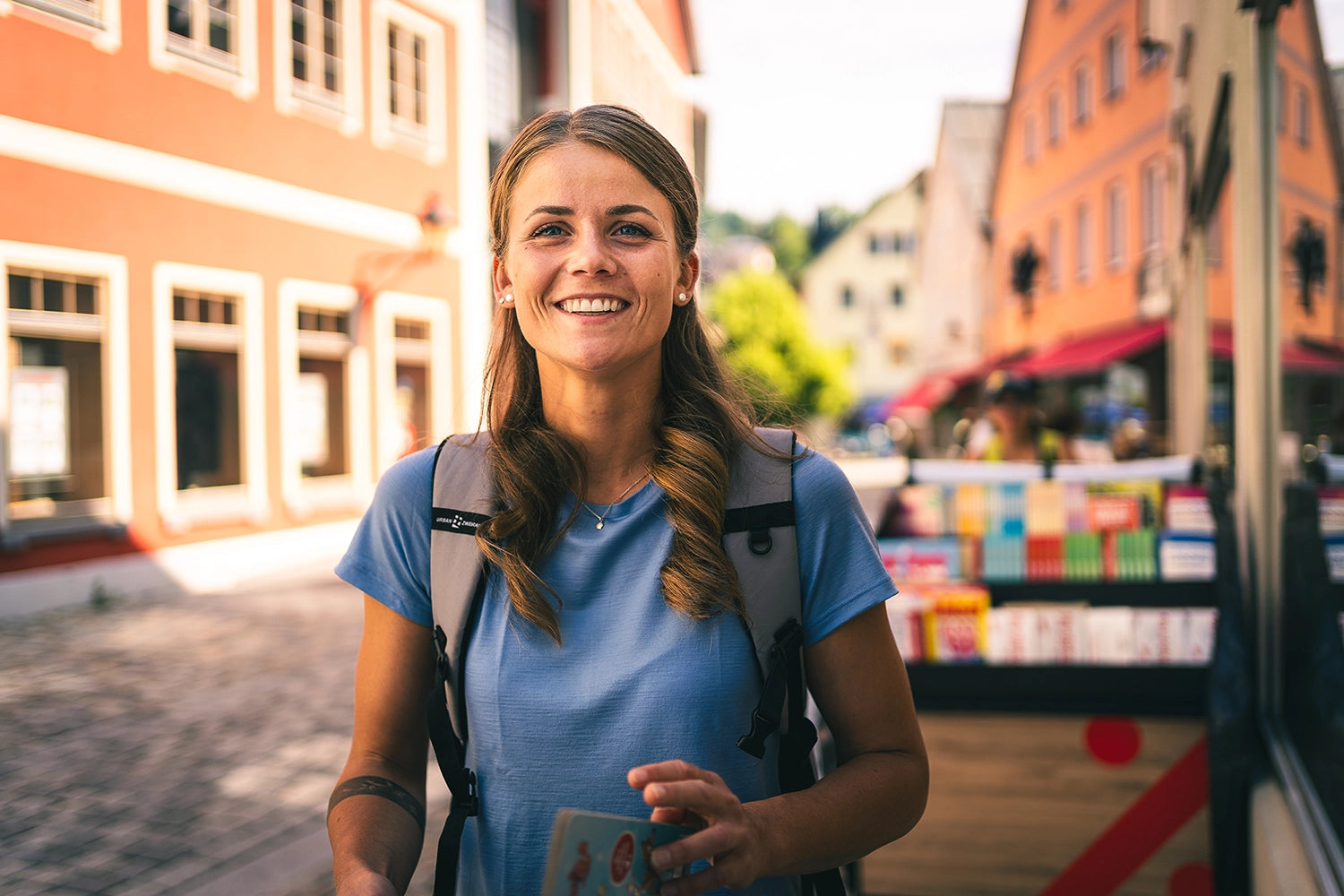 Frau trägt ein blaues TOM FYFE Merino-Shirt mit Rucksack in einer sonnigen Altstadt.