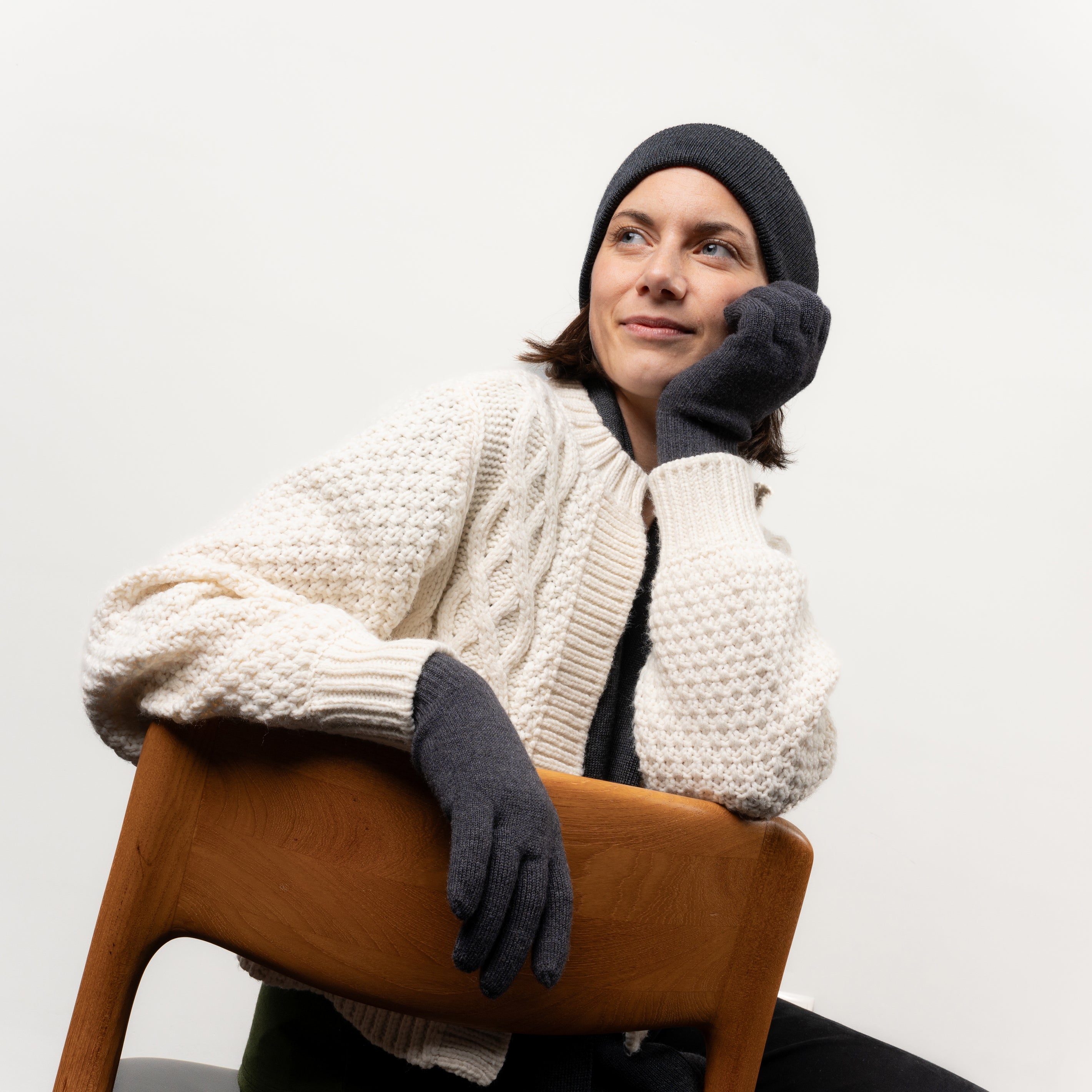 Frau mit anthrazitfarbenen Strickhandschuhen und einer Beanie aus Merinwolle sitzt gemütlich auf einem Stuhl
