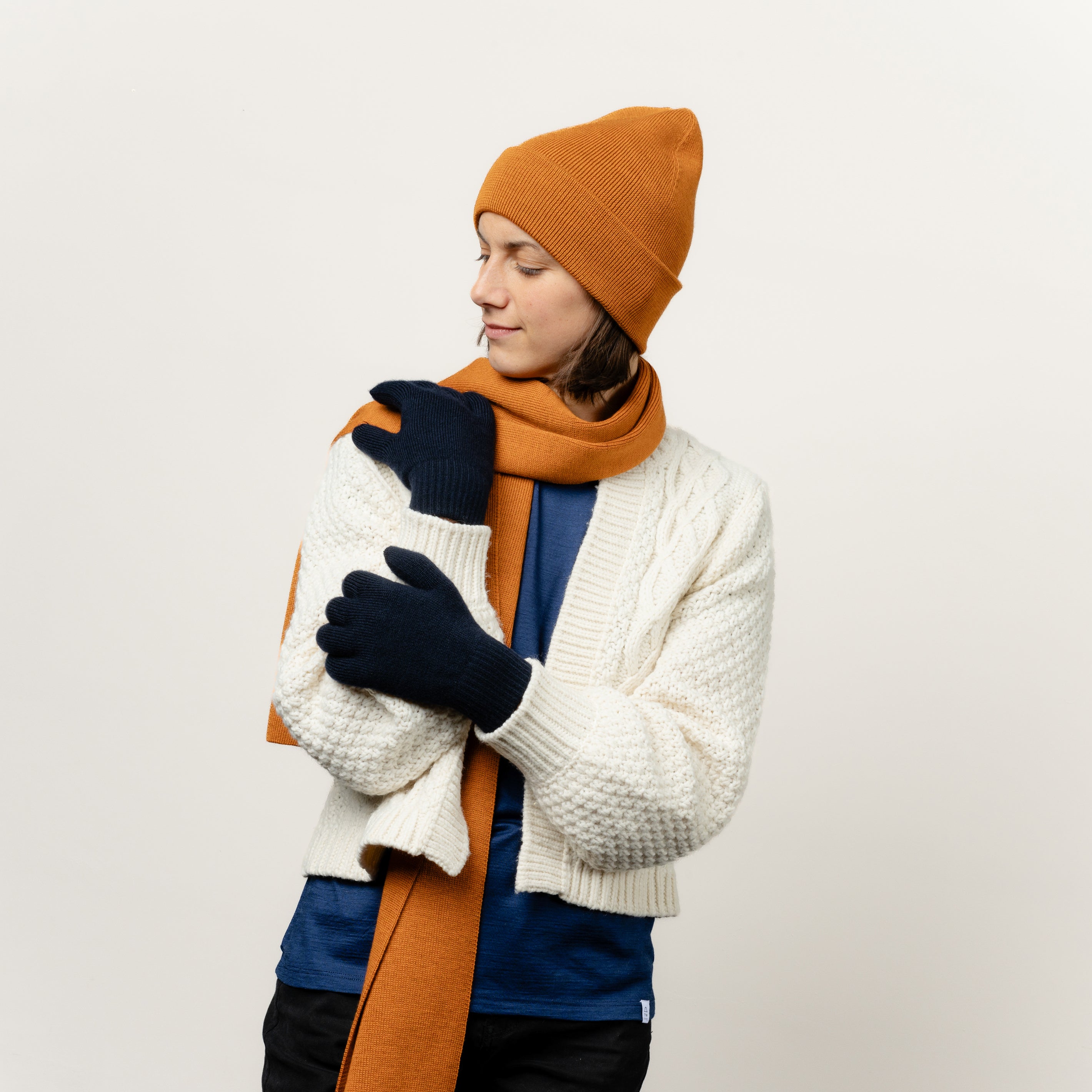 Weibliches Model kombinierte ihre Wollhandschuhe in der Farbe Dunkelblau mit einem ockerfarbenen Schal aus Merinwolle und einer gleichfarbigen Mütze von Tom Fyfe