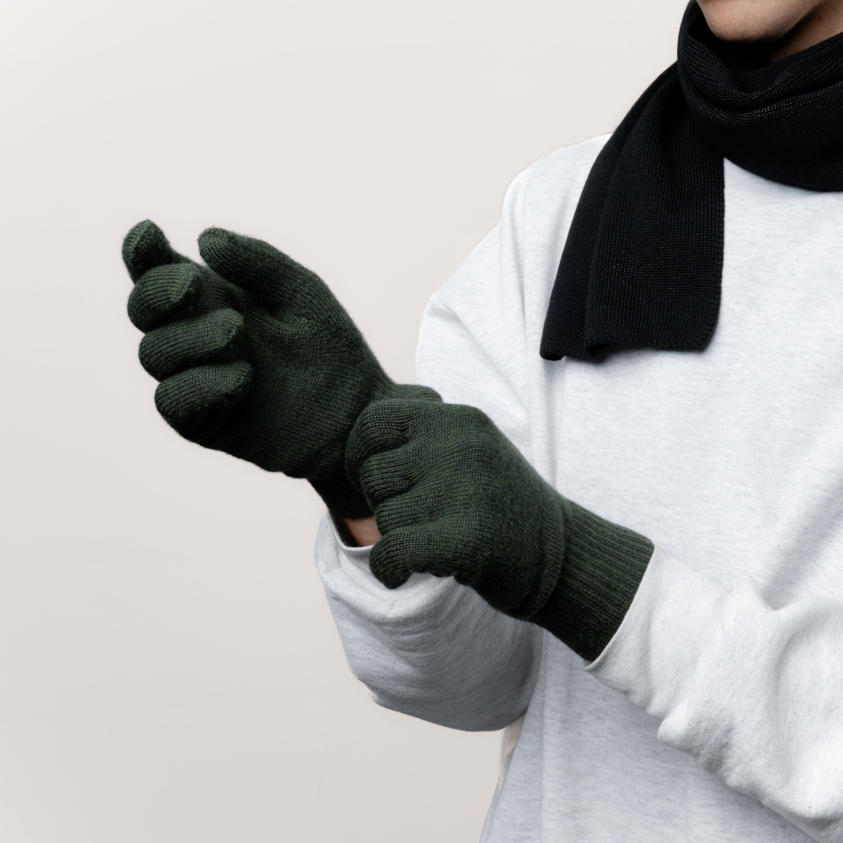 Männliches Model zieht seine Strickhandschuhe aus Merinowolle von Tom Fyfe in der Farbe Dunkelgrün an. Dazu trägt es einen Schal aus Merinowolle von Tom Fyfe