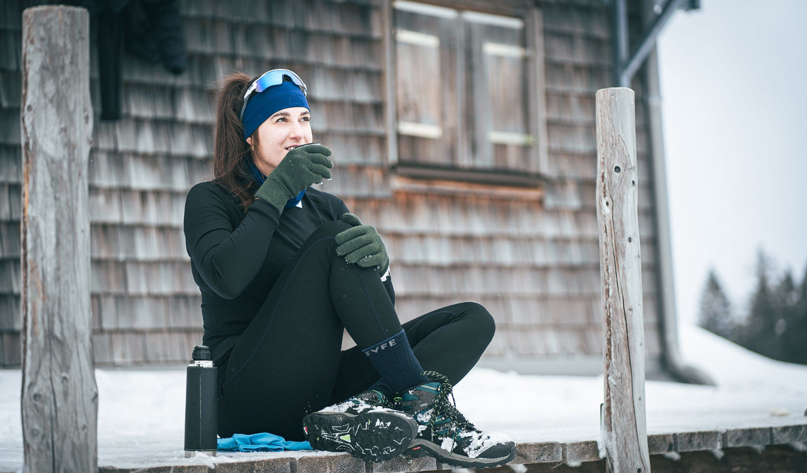 Frau in TOM FYFE Activewear sitzt vor einer verschneiten Holzhütte und trinkt Tee