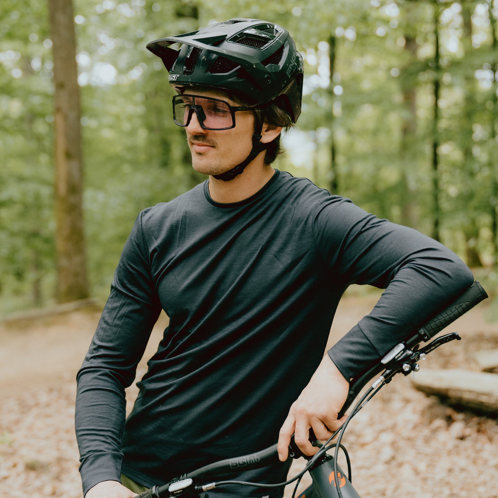 Männlicher Fahrradfahrer steht mit Helm und Brille im Wald. Er trägt ein Merino-Longlseeve von Tom Fyfe.