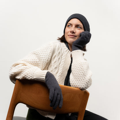 Frau mit anthrazitfarbenen Strickhandschuhen und einer Beanie aus Merinwolle sitzt gemütlich auf einem Stuhl 