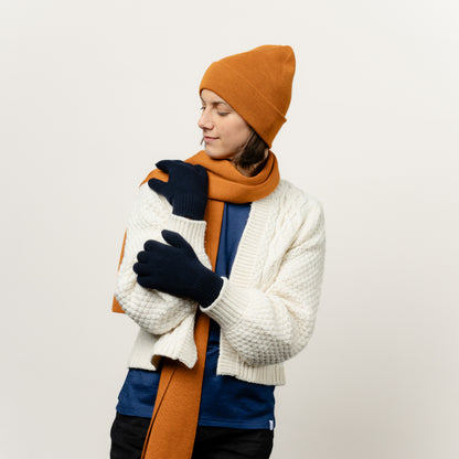 Weibliches Model kombinierte ihre Wollhandschuhe in der Farbe Dunkelblau mit einem ockerfarbenen Schal aus Merinwolle und einer gleichfarbigen Mütze von Tom Fyfe 
