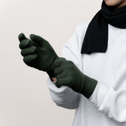 Männliches Model zieht seine Strickhandschuhe aus Merinowolle von Tom Fyfe in der Farbe Dunkelgrün an. Dazu trägt es einen Schal aus Merinowolle von Tom Fyfe 