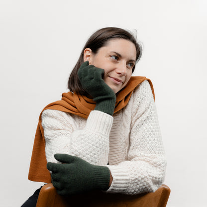 Weibliches Model kombinierte ihre Strickhandschuhe in Dunkelgrün mit einem Schal in der Farbe Ocker von Tom Fyfe und sitzt auf einem Stuhl 